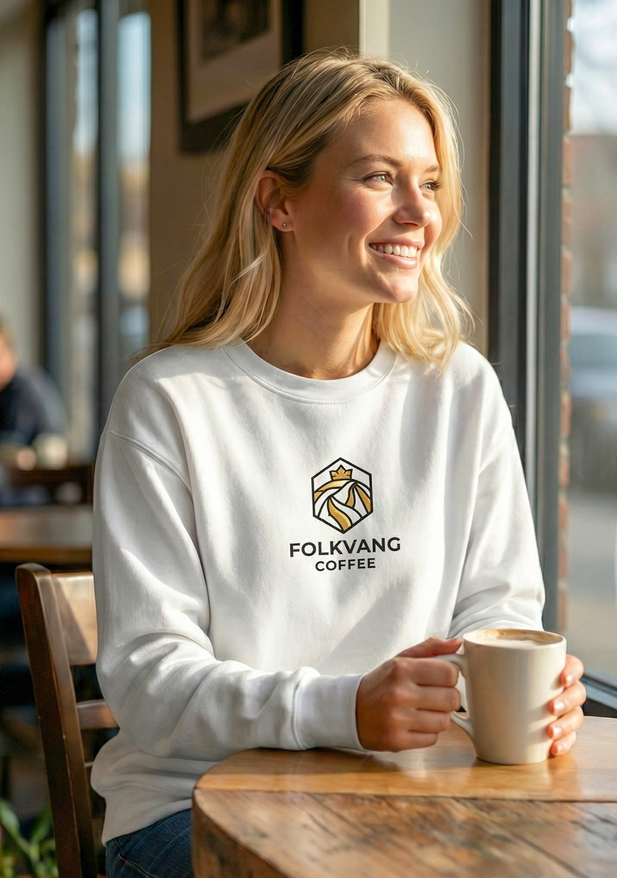 Woman wearing a white sweatshirt with Folkvang Coffee logo, holding a coffee mug indoors.